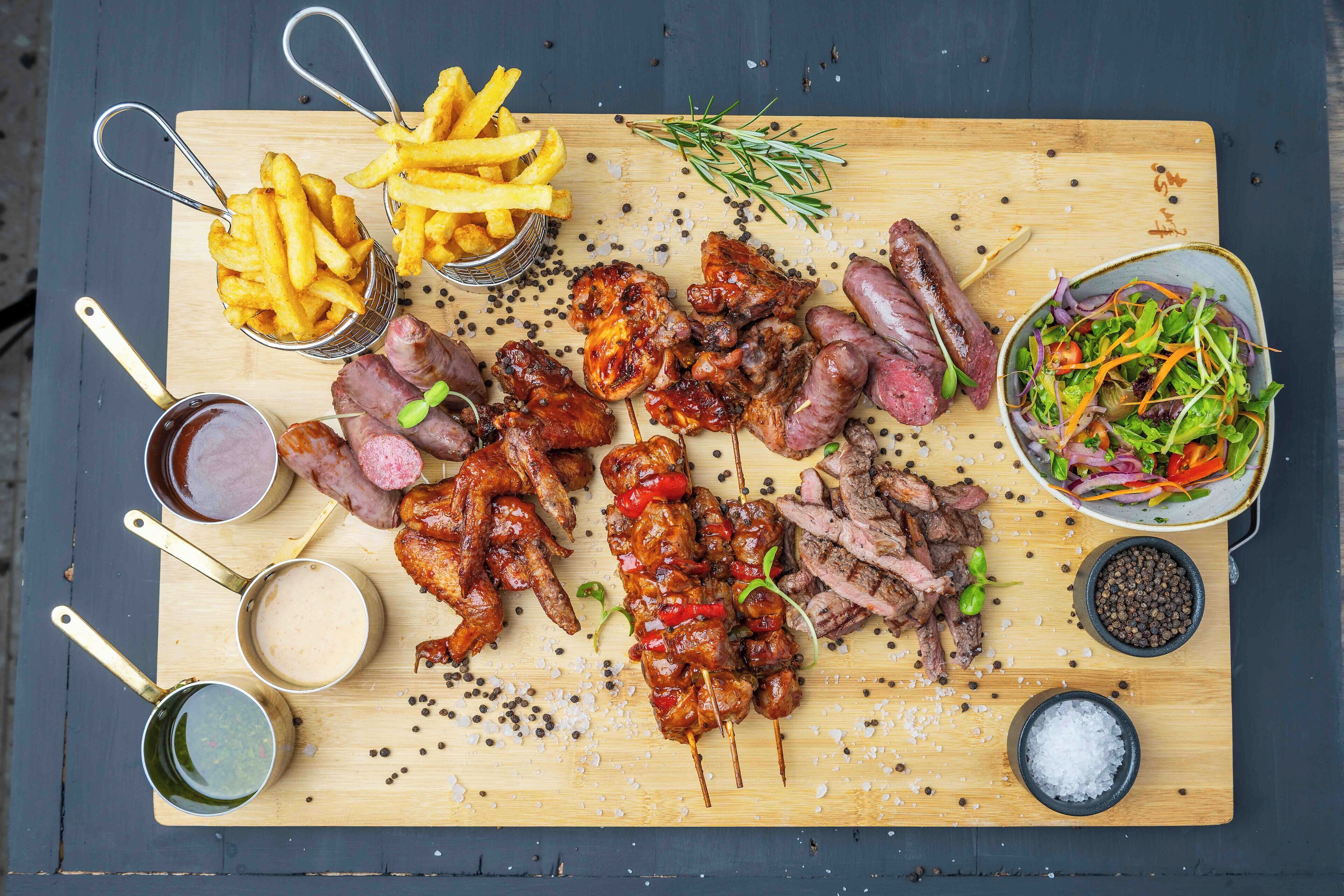 A wooden board with various meats, fries, and a salad.