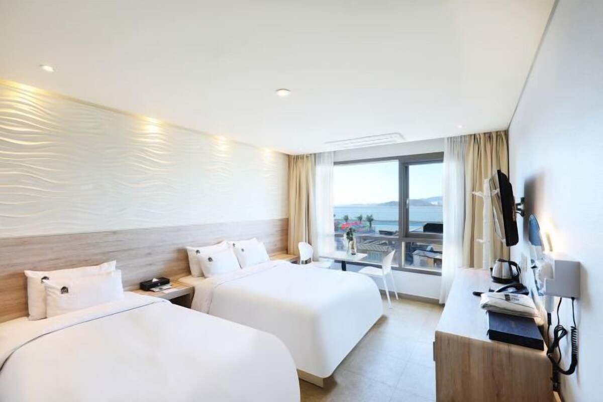 A hotel room with two beds, a desk with a computer, and a large window offering a view of the sea.