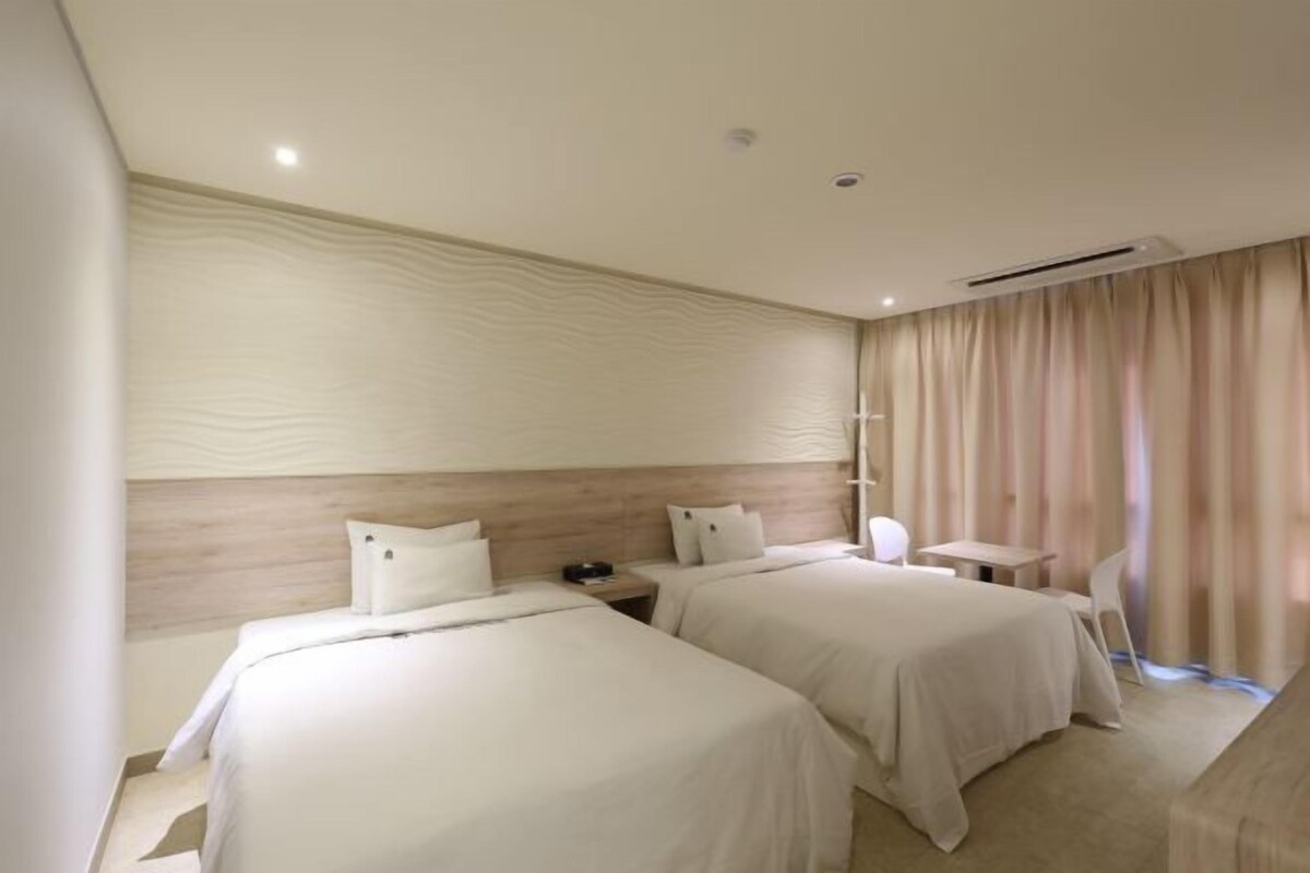 A hotel room with two beds, a small table, a chair, and a window with curtains.