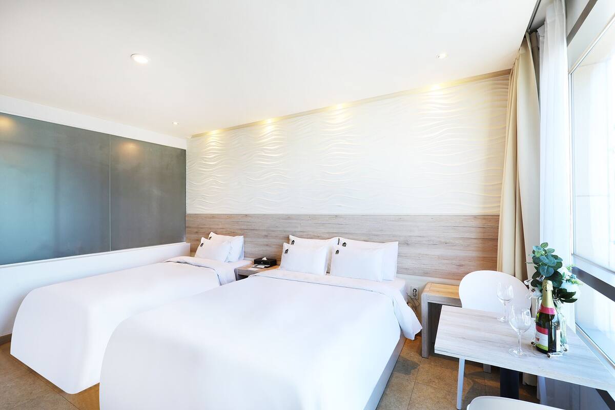 A modern hotel room with two beds, a small table with wine glasses, and a view of the city.
