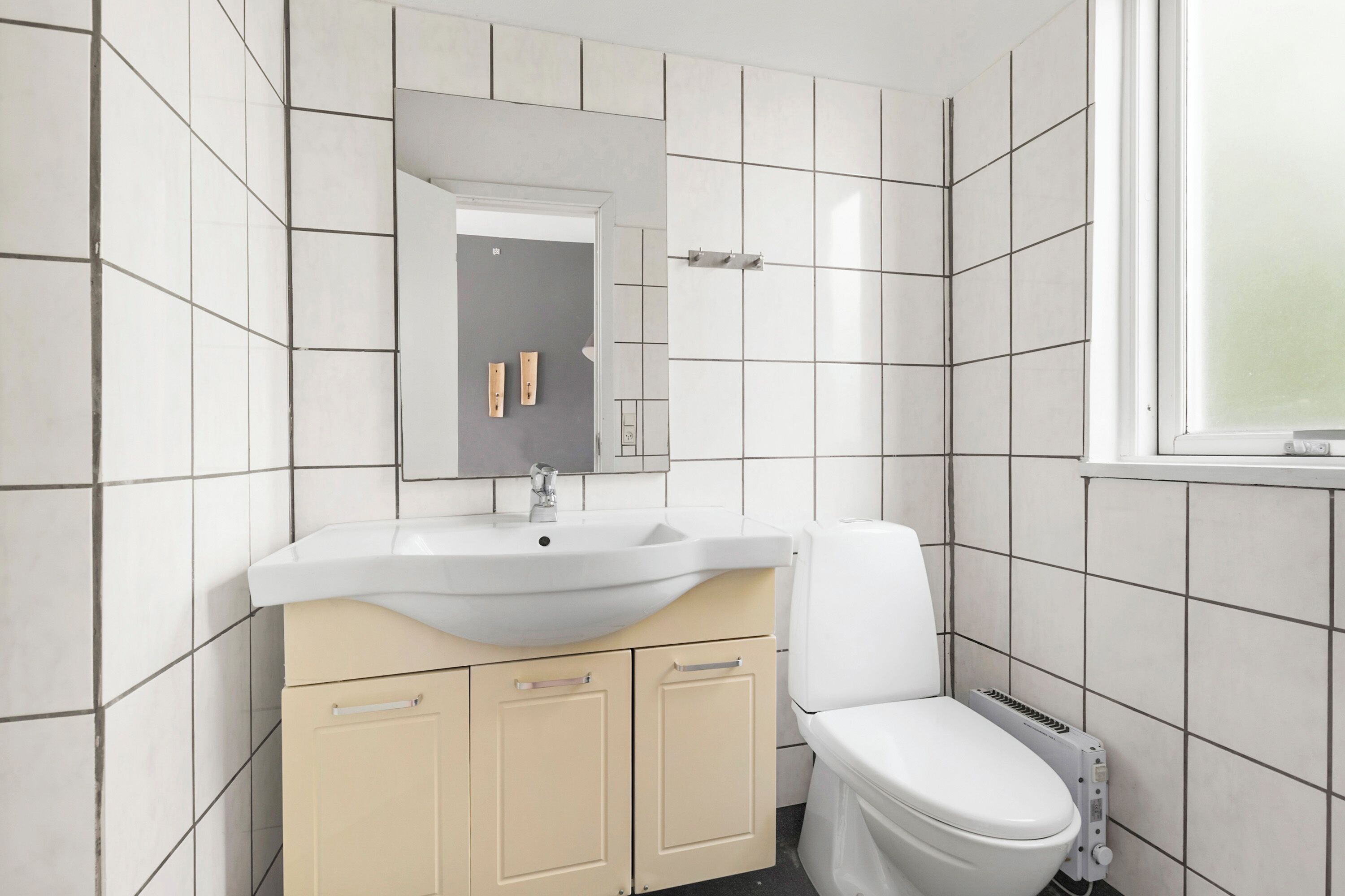 Un baño con azulejos blancos, un lavamanos color beige, un inodoro blanco y una ventana.