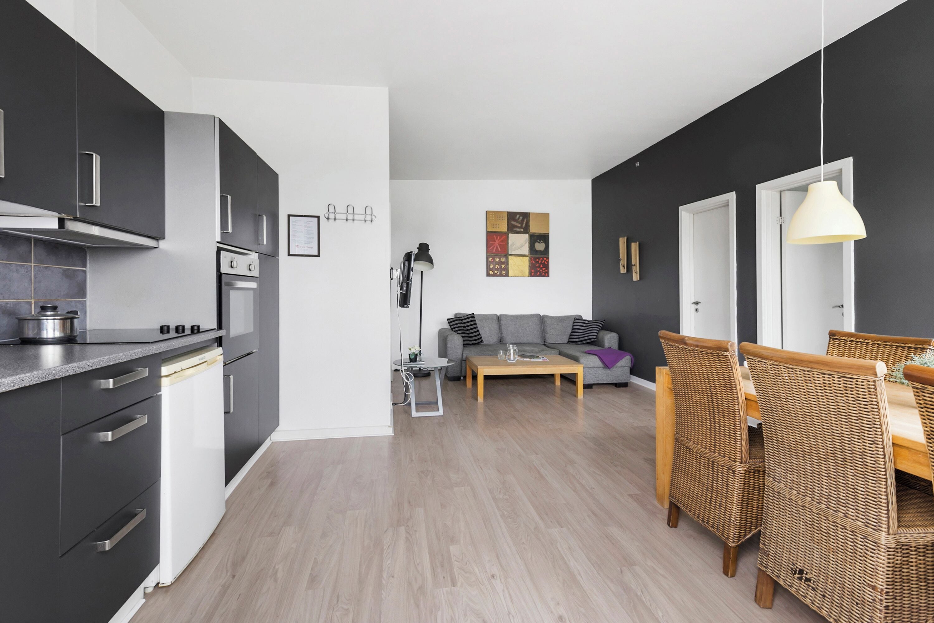 A modern kitchen with dark cabinets, a built-in oven, and a dishwasher. There is a dining area with wicker chairs and a wooden table.
