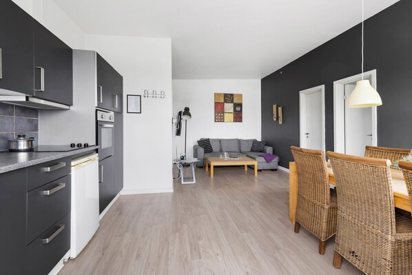 A modern kitchen with dark cabinets, a built-in oven, and a dishwasher. There is a dining area with wicker chairs and a wooden table. - First Camp Tempelkrogen - Holbæk (Vipperod)