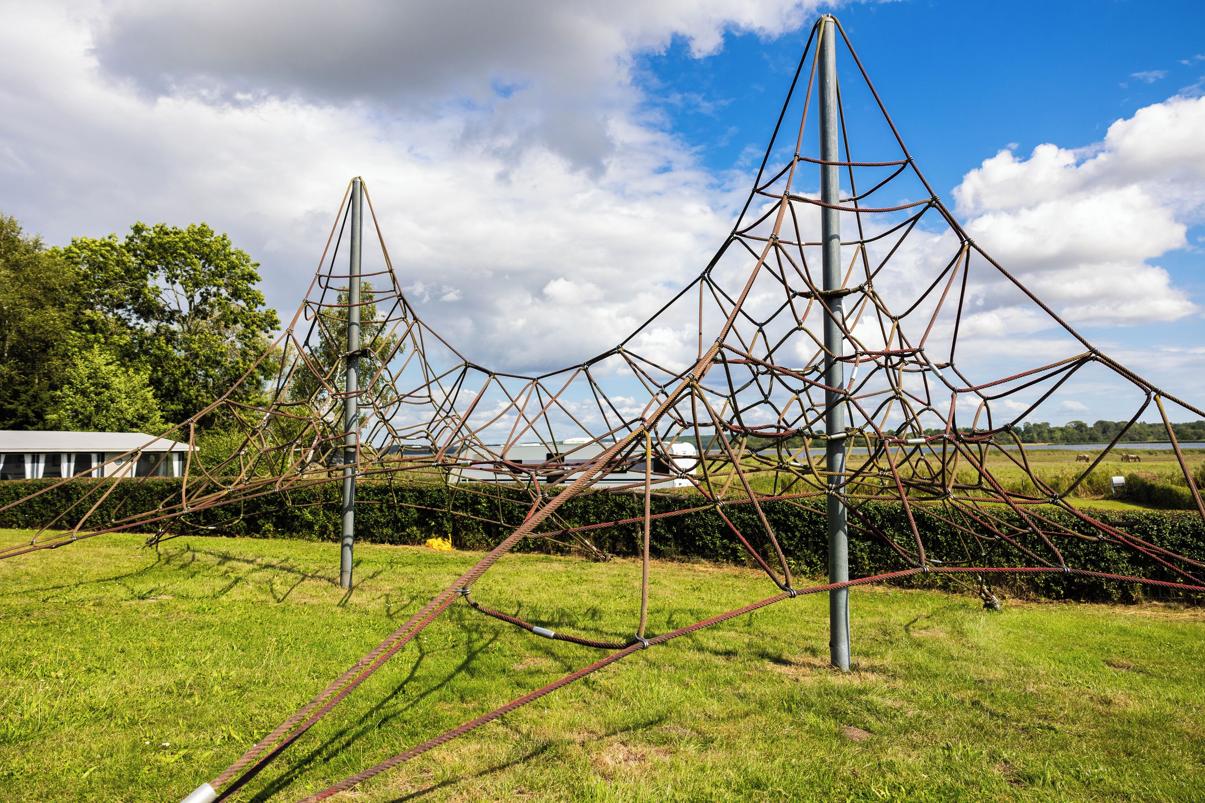 Una gran estructura metálica con una intrincada red de cables y postes, ubicada en un campo cubierto de hierba con árboles y un edificio al fondo.