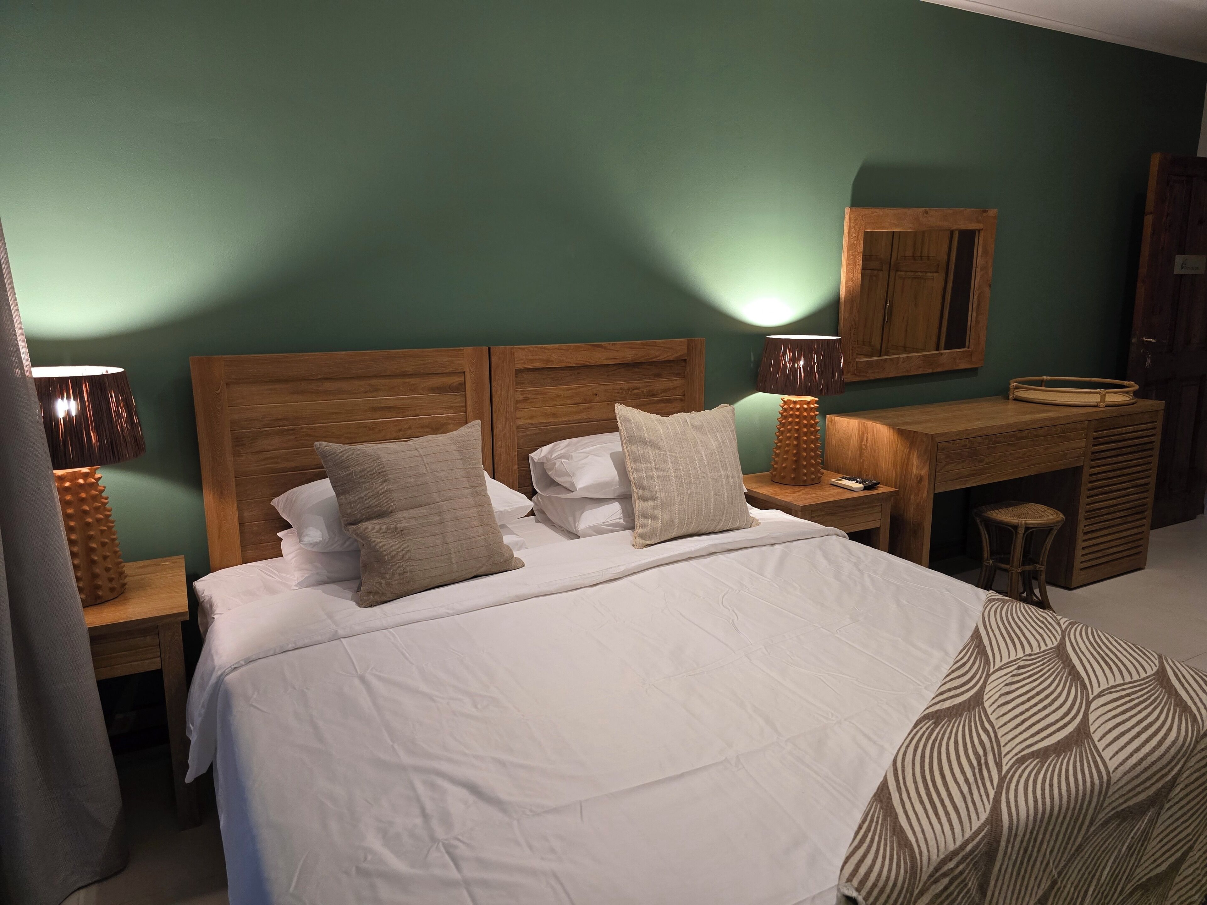 A neatly made bed with two pillows, a wooden nightstand with a lamp, a small wooden desk with a chair, and a wall-mounted cabinet.
