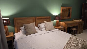 Una cama bien hecha con dos almohadas, una mesita de noche de madera con una lámpara, un escritorio pequeño de madera y una silla, y un mueble de pared.