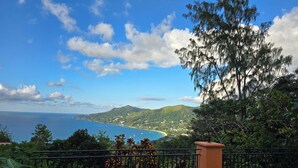 Paisaje costero con vista despejada al mar, colinas y un jardín cercado con plantas tropicales.