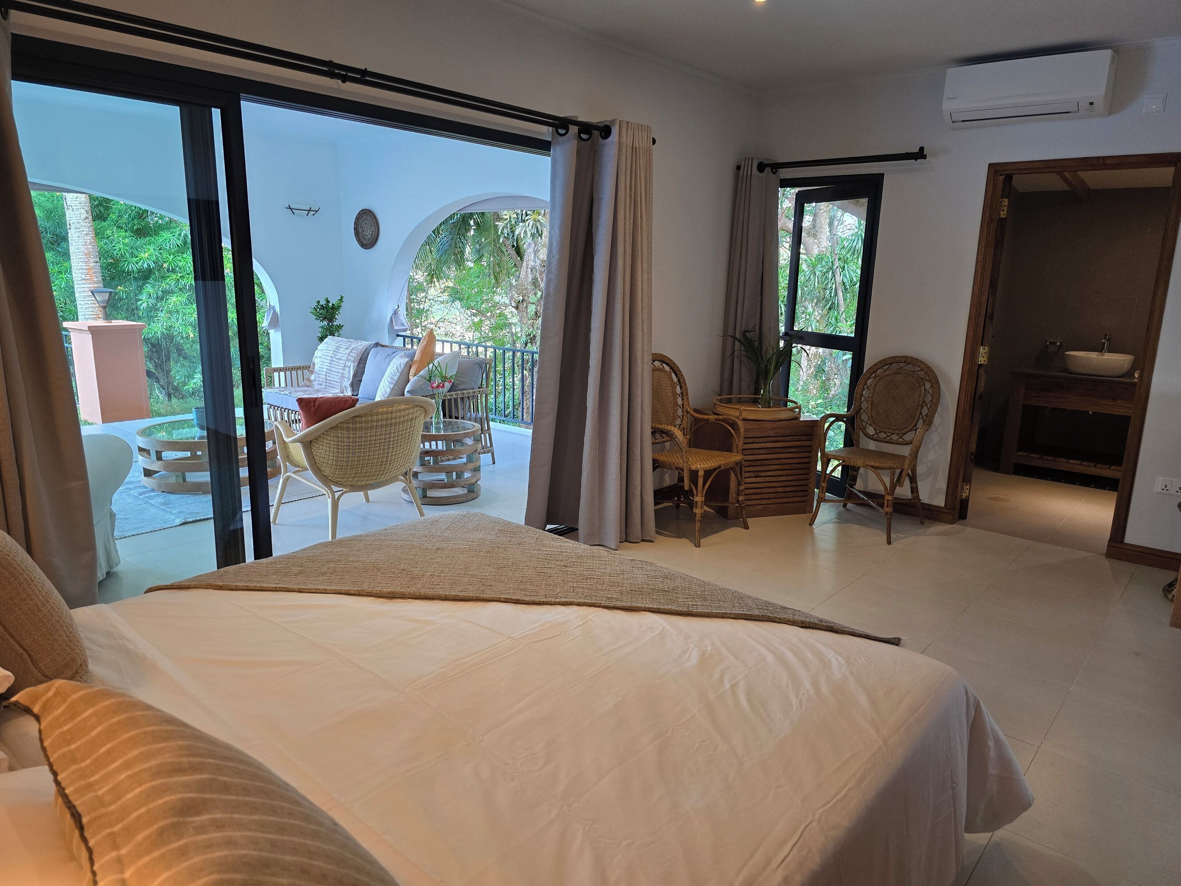 A hotel room with a large bed, a balcony with wicker furniture, and a view of greenery.