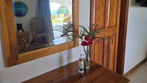Un espejo con marco de madera que refleja una habitación con una cafetera, una silla y una mesa con una botella y un florero con plantas.