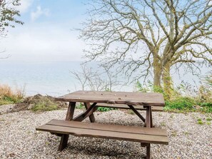 A wooden picnic table with benches, set on a gravel area with a view of a body of water and leafless trees in the background.