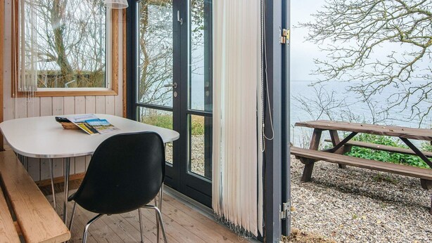 A room with a table and chair, a bench outside, and a view of the sea through large windows.