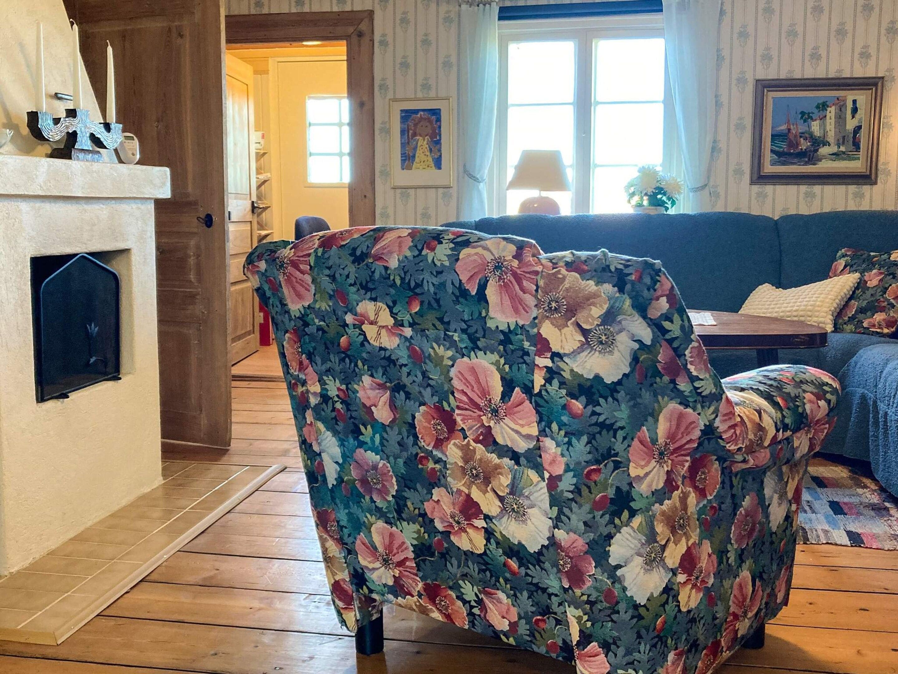 A living room with a floral-patterned armchair, a blue sofa, a fireplace, and a window with curtains.