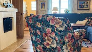 A living room with a floral-patterned armchair, a blue sofa, a fireplace, and a window with curtains.