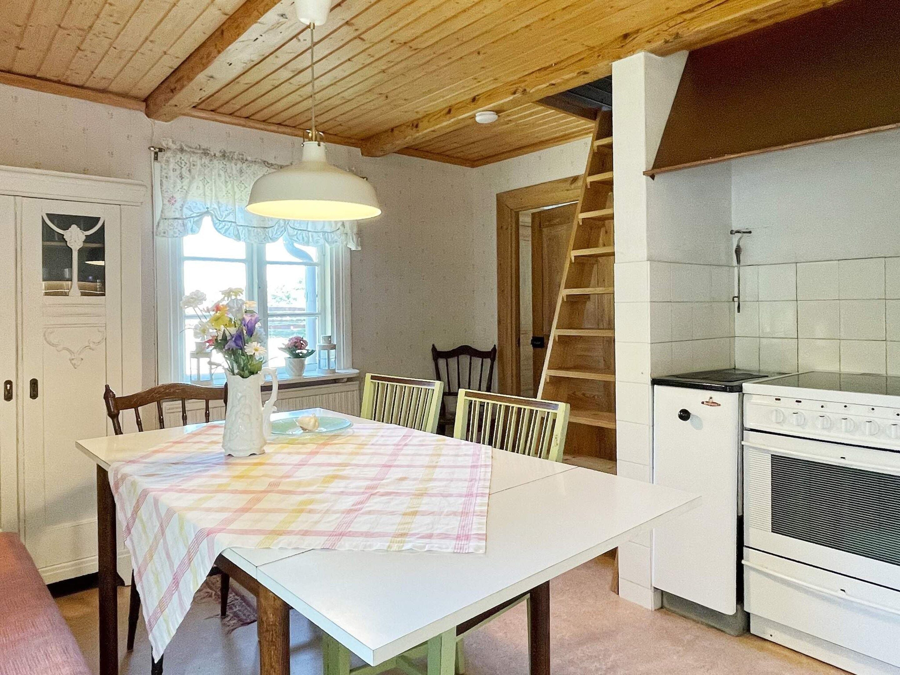 Un comedor con una mesa lista para dos, una ventana con cortinas, techo de madera y una zona de cocina con estufa y armarios.