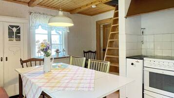 Un comedor con una mesa lista para dos, una ventana con cortinas, techo de madera y una zona de cocina con estufa y armarios.