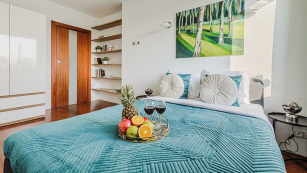 Un dormitorio con una cama, una cesta de frutas, dos copas de vino y un cuadro en la pared.