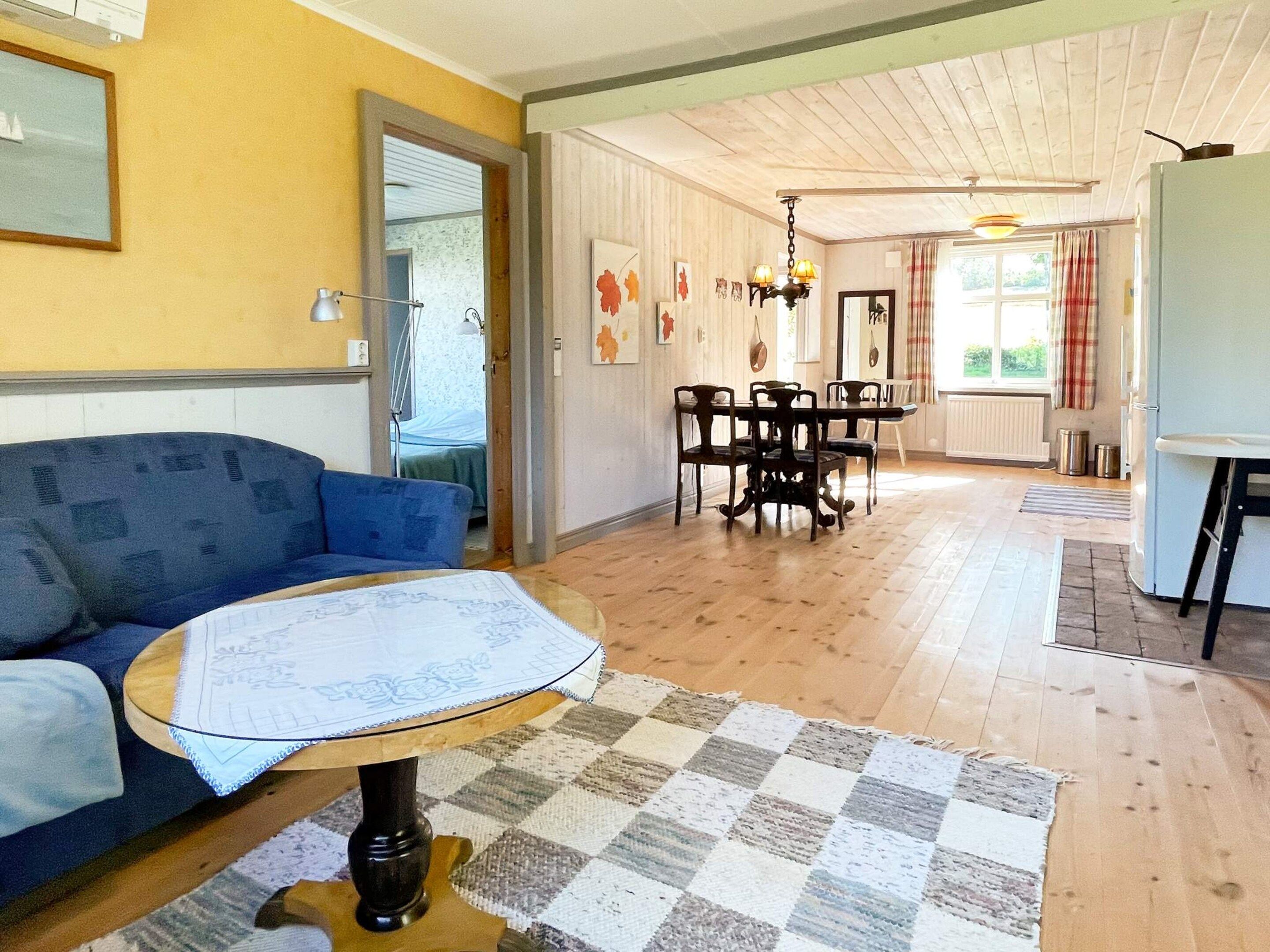 Una habitación con un sofá azul, una mesa redonda con mantel de encaje, un tapete a cuadros, una mesa de comedor con sillas y un refrigerador blanco.