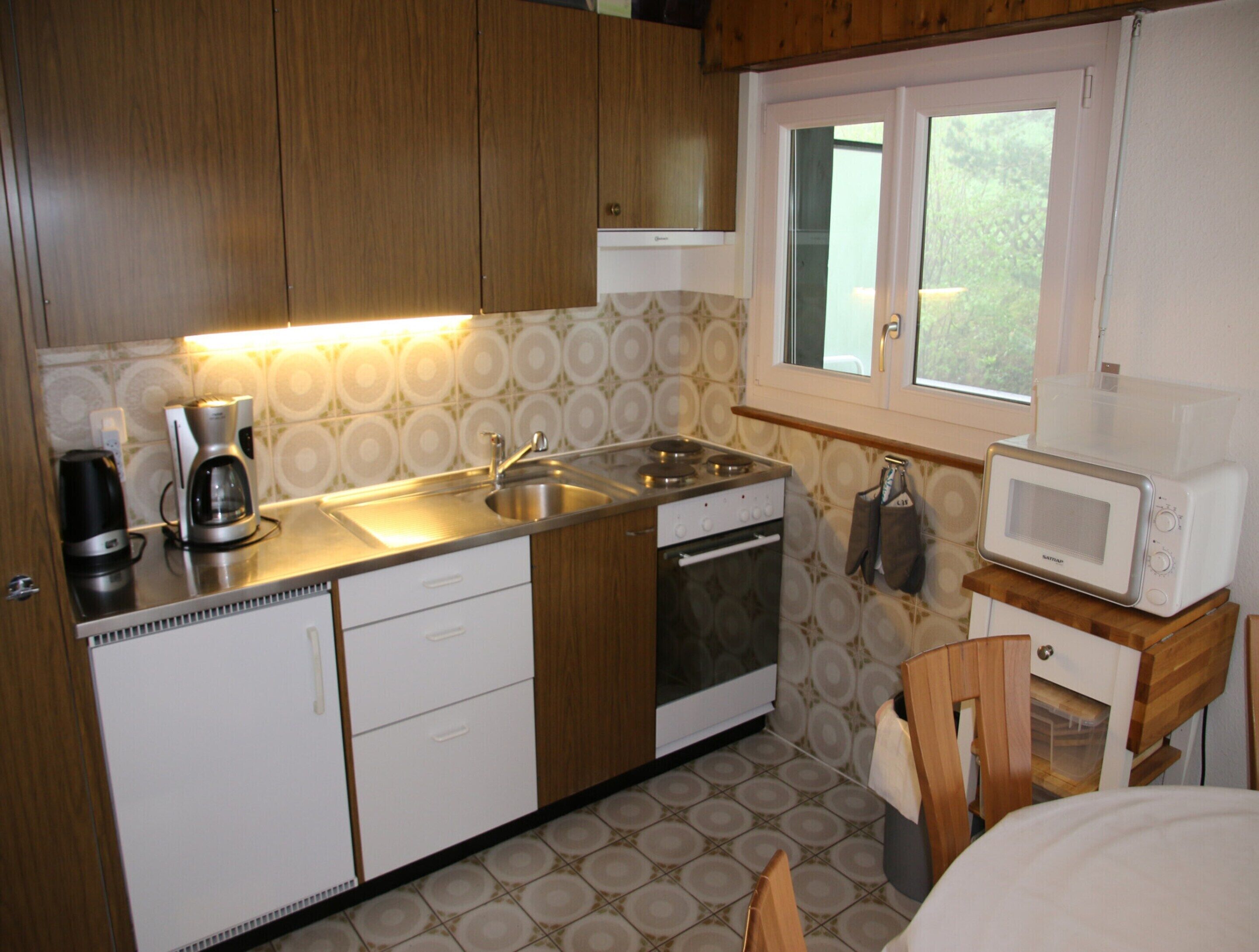 A compact kitchen with wooden cabinets, a stainless steel sink, a built-in oven, and a microwave on a wooden stand.