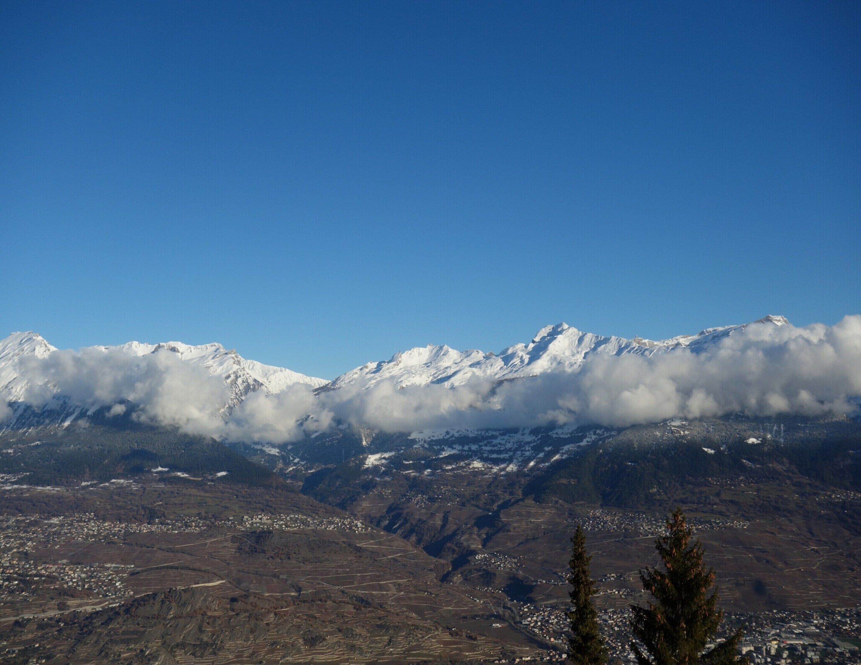 A mountainous landscape with snow-capped peaks, a valley with scattered buildings, and a clear blue sky.