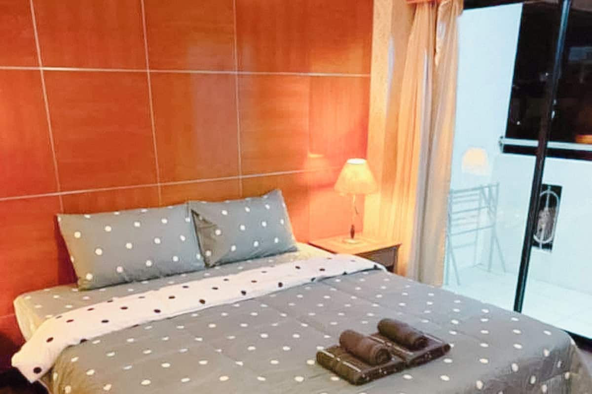 A hotel room with a wooden headboard, a bed with a polka dot bedspread, a small bedside table with a lamp, and a view of the outside through a large window.