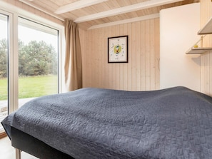 A bedroom with a large bed, a window with curtains, a framed picture on the wall, and a shelf with a lamp.