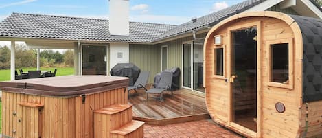 Et træspabad og sauna tilknyttet et hus med en overdækket terrasse og udendørs møbler.