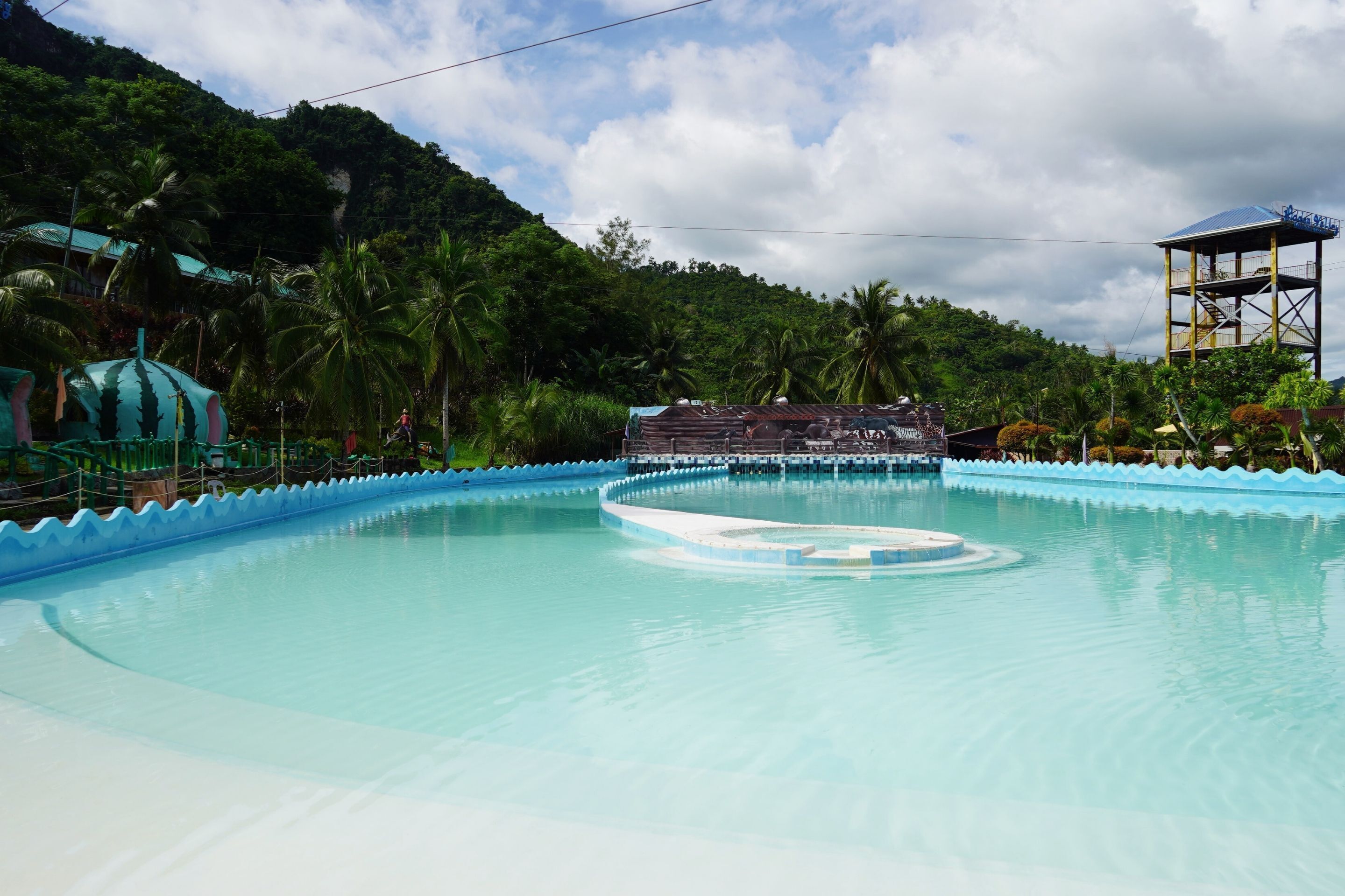 A large, clear pool with a blue edge, surrounded by lush greenery and a hill in the background.