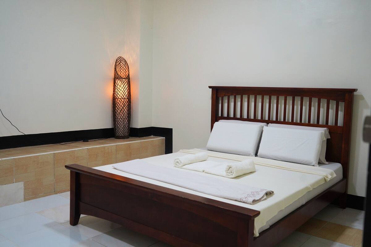 Una cama bien hecha con ropa blanca y toallas, cabecera de madera y una lámpara fijada a la pared.