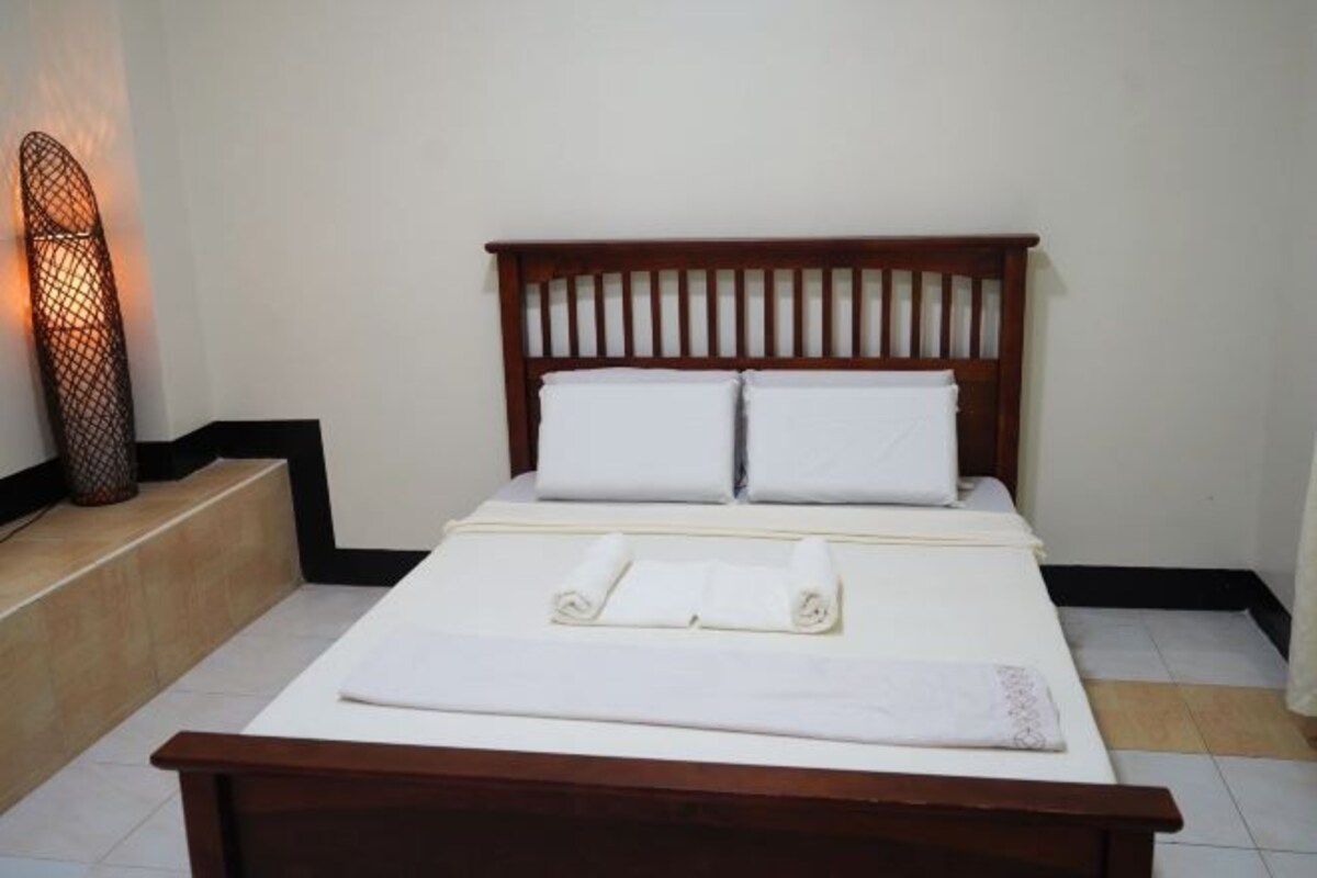 A neatly made bed with white linens and pillows, a wooden headboard, and a bedside lamp.