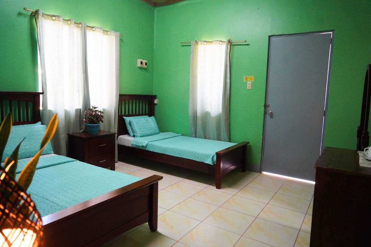 Una habitación con dos camas, un aparador de madera, una puerta y un ventanal con cortinas.