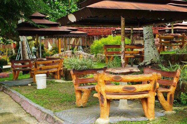 Des bancs et des tables en bois, installés à l’extérieur, sous des toits en métal rouillé, le tout entouré de verdure.
