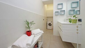 Ein modernes Badezimmer mit einem weißen Waschbecken, einem Spiegel mit blauen Regalen, einer Waschmaschine und einer Sitzbank mit Handtüchern.