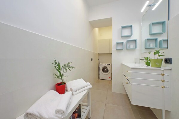 A modern bathroom with a white sink, a mirror with blue shelves, a washing machine, and a bench with towels. - Luminoso appartamento a Testaccio (Rome)