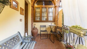 Un patio con un muro de ladrillo, una ventana de madera, una mesa de metal, dos sillas de metal, un banco de madera y plantas en macetas.