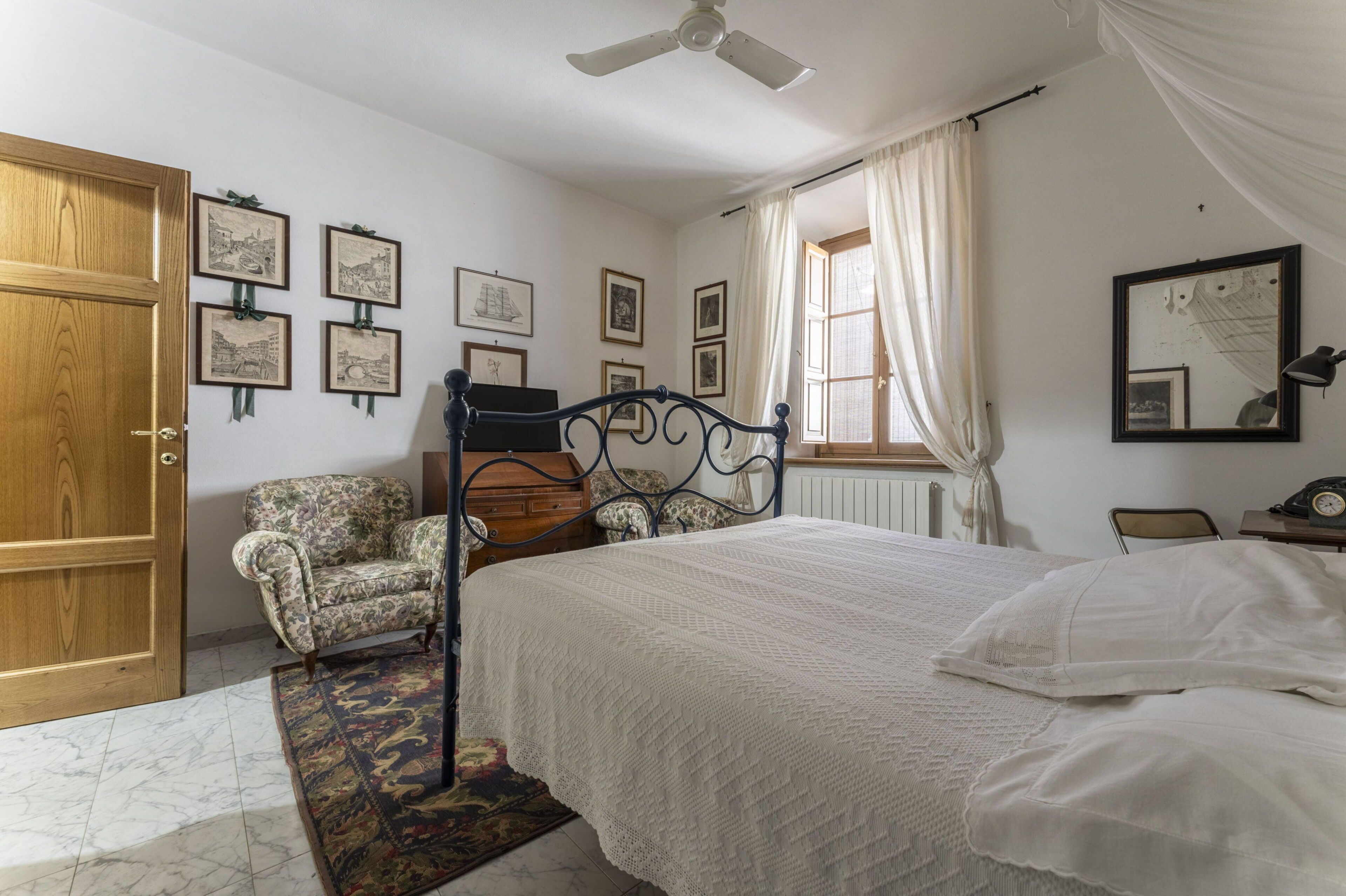 A bedroom with a bed, a chair, a wooden wardrobe, a ceiling fan, and framed pictures on the wall.