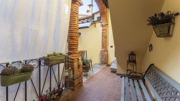 Un estrecho pasillo con columnas de ladrillo, un banco y macetas colgantes con flores.