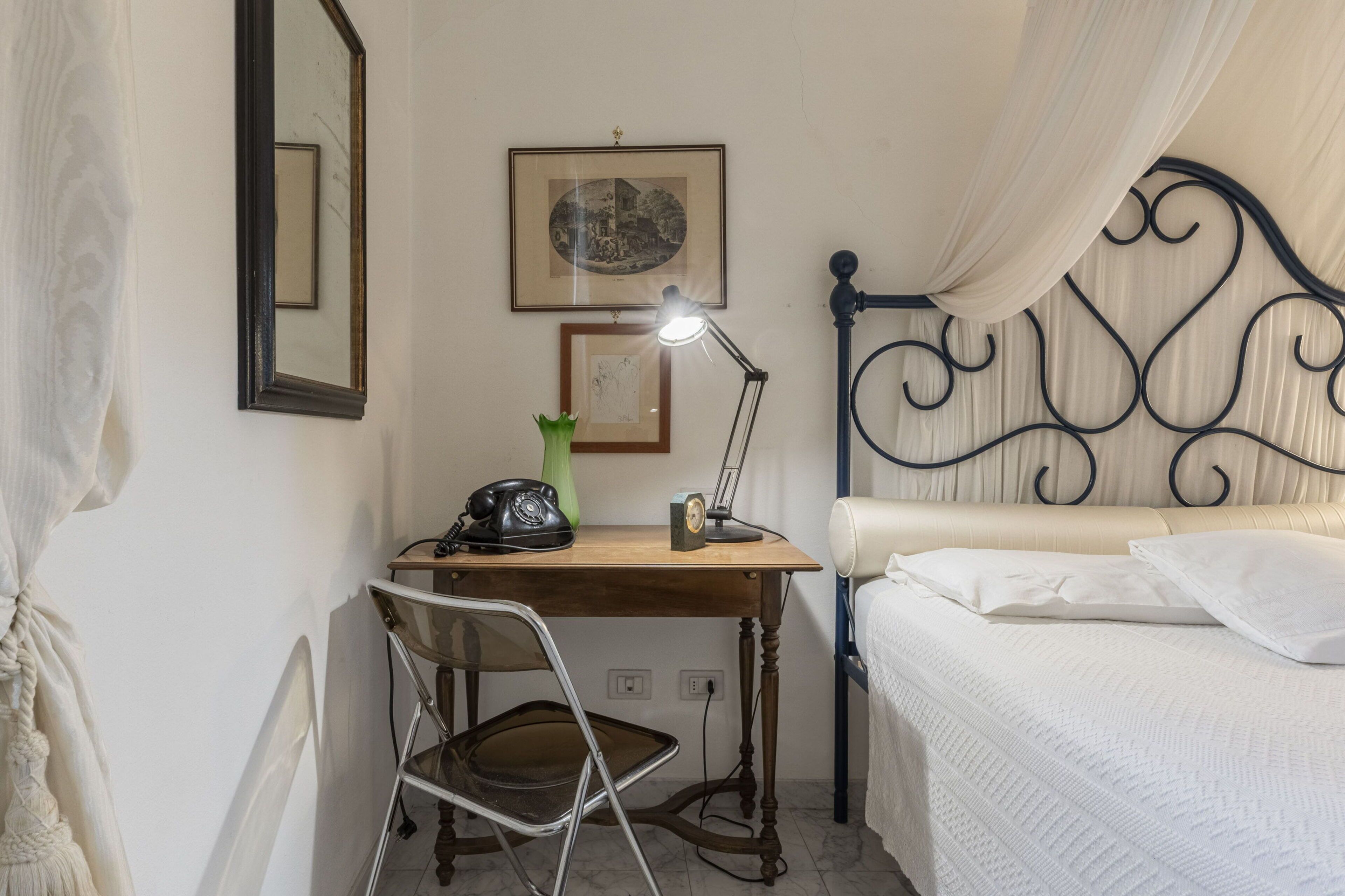 Een slaapkamer met een bed, een bureau met een lamp, een stoel en een klein tafeltje met een telefoon en een vaas.