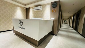 Ein Empfangsschalter mit einem Kronenlogo und dem Schriftzug "SHREE JI INN" in einer Hotelhalle mit gemustertem Wandpapier, einer Uhr und einem Schild, das Ein- und Auscheckzeiten angibt.