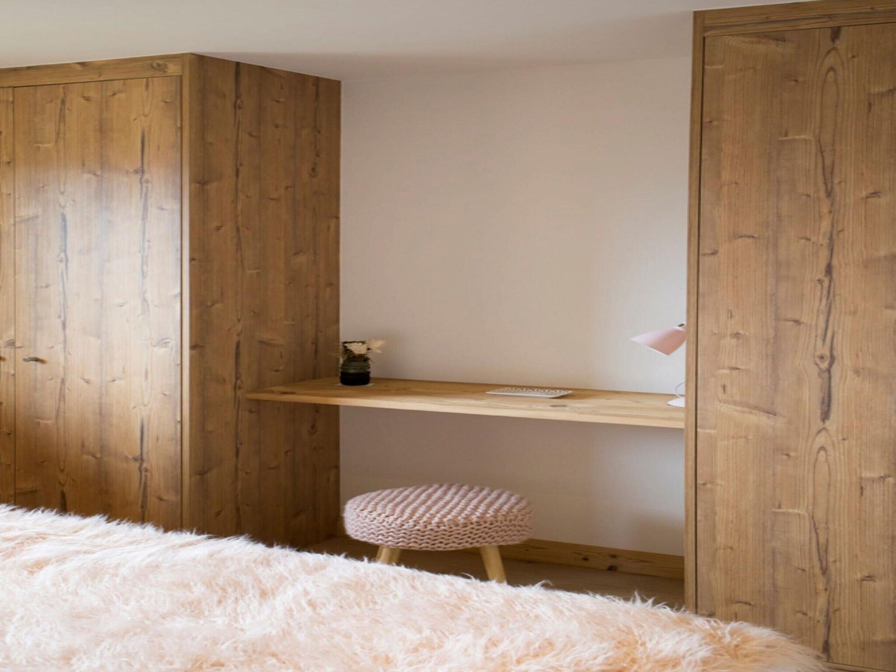 A bedroom with a wooden wardrobe, a small desk with a lamp, a fluffy rug, and a round stool.