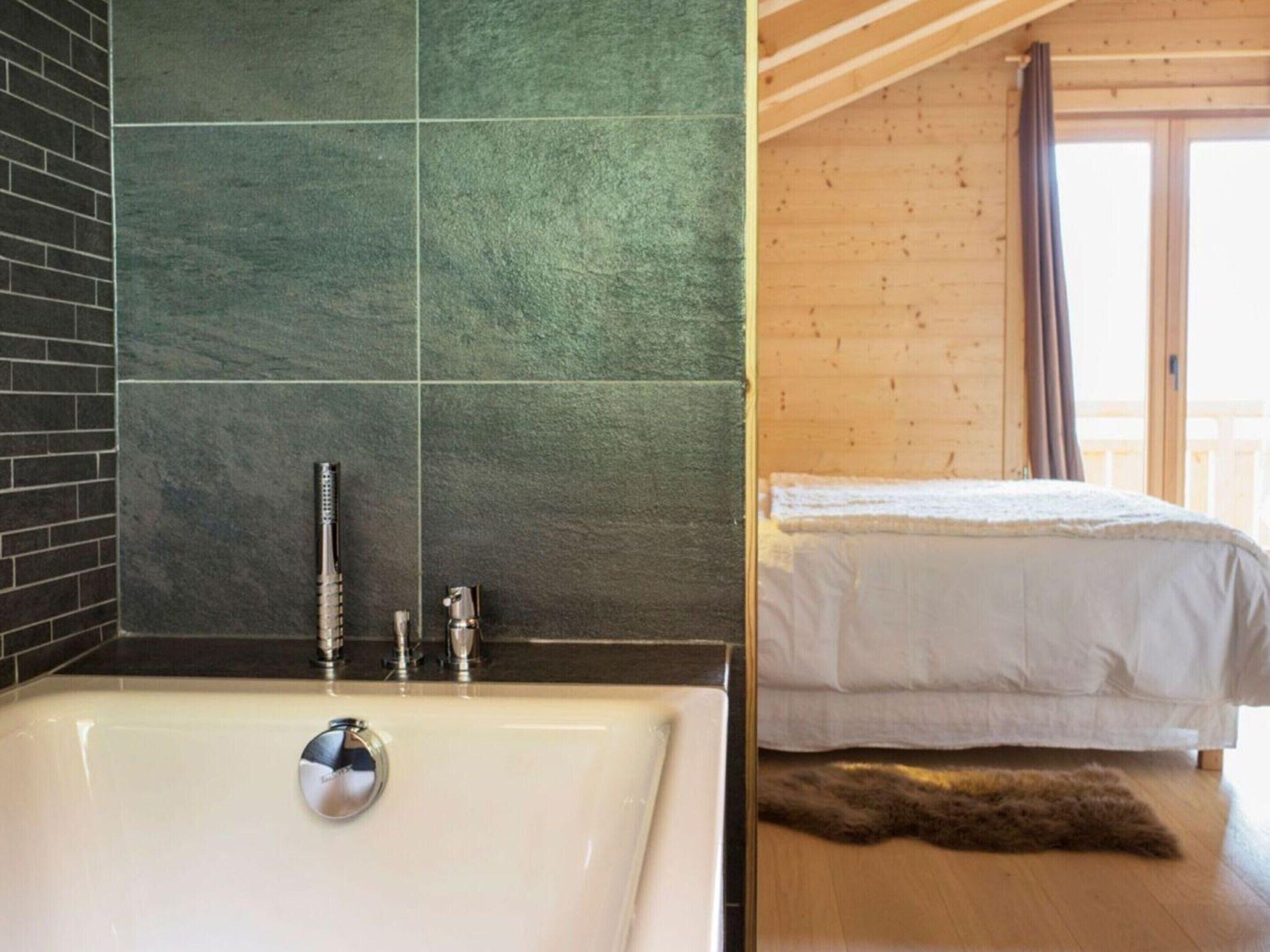 A bathroom with a bathtub, a shower, and a sink. There is a bed with a white blanket and a furry rug on the floor.