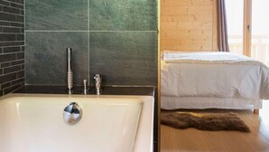 Une salle de bain avec une baignoire, une douche et un lavabo. On y trouve un lit recouvert d’une couverture blanche et un tapis moelleux sur le sol.