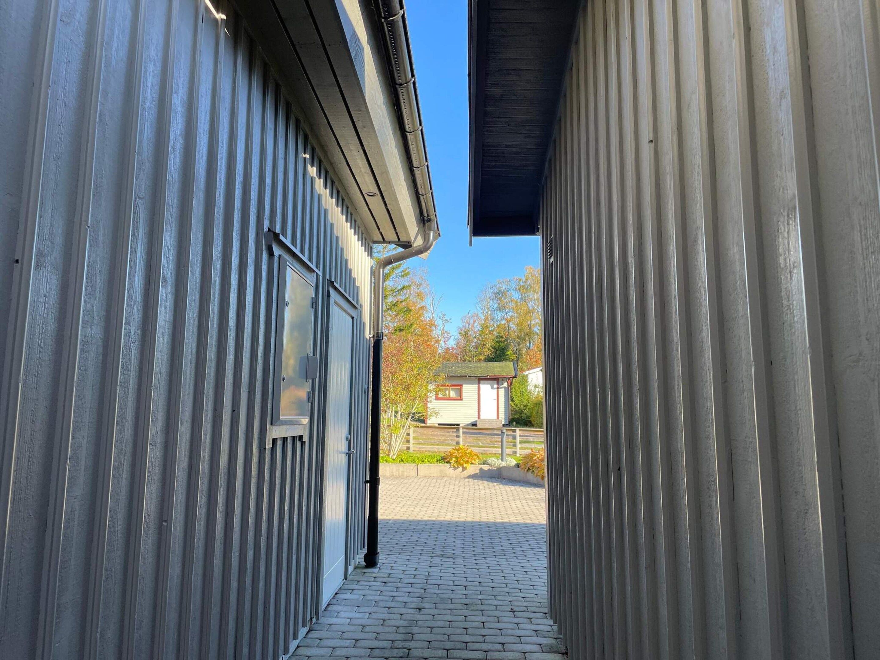 A narrow corridor with metal walls, a brick pathway, and a small building in the distance.