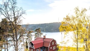 A red house with a sloped roof, a chimney, and a small porch with a blue door. It is surrounded by trees and a body of water. - 4 Star Holiday Home in Valdemarsvik (Valdemarsvik)