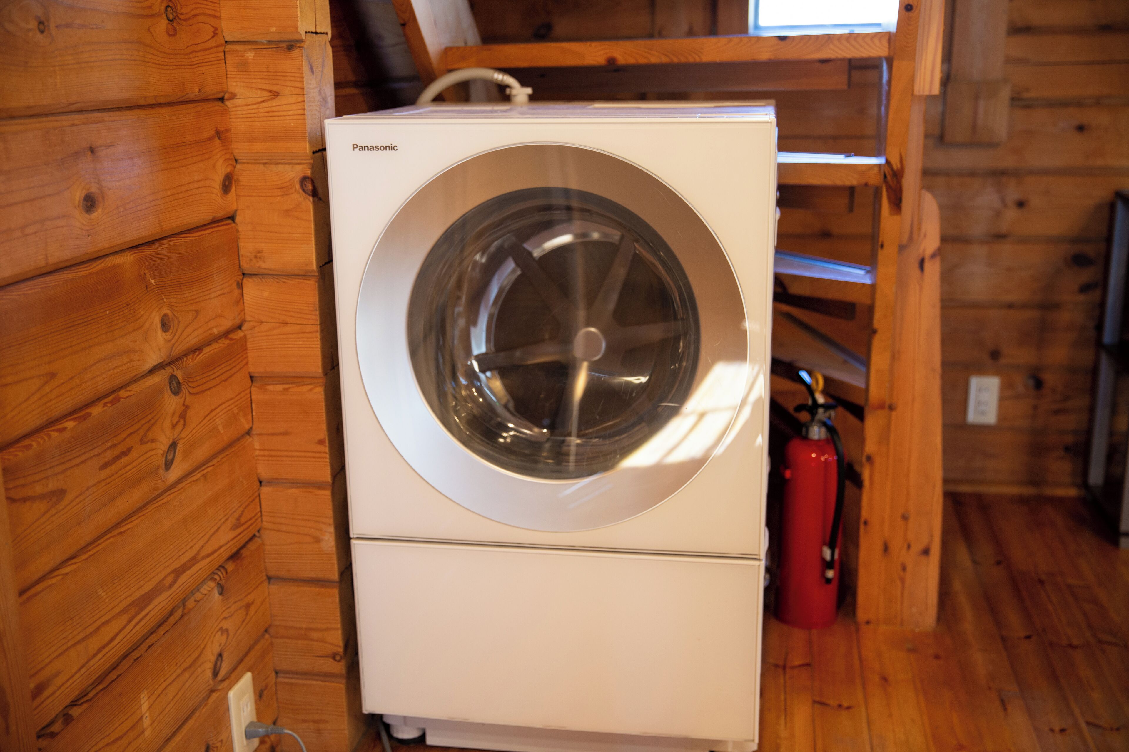 Een witte wasmachine in een houten chalet, met een brandblusser in de buurt.