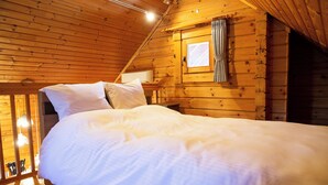 Une chambre dans un chalet en bois, avec un lit, des oreillers, une fenêtre avec des rideaux et une petite table.