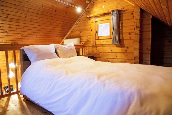A wooden cabin bedroom with a bed, pillows, a window with curtains, and a small table. - Three Peaks Log (Hakuba)
