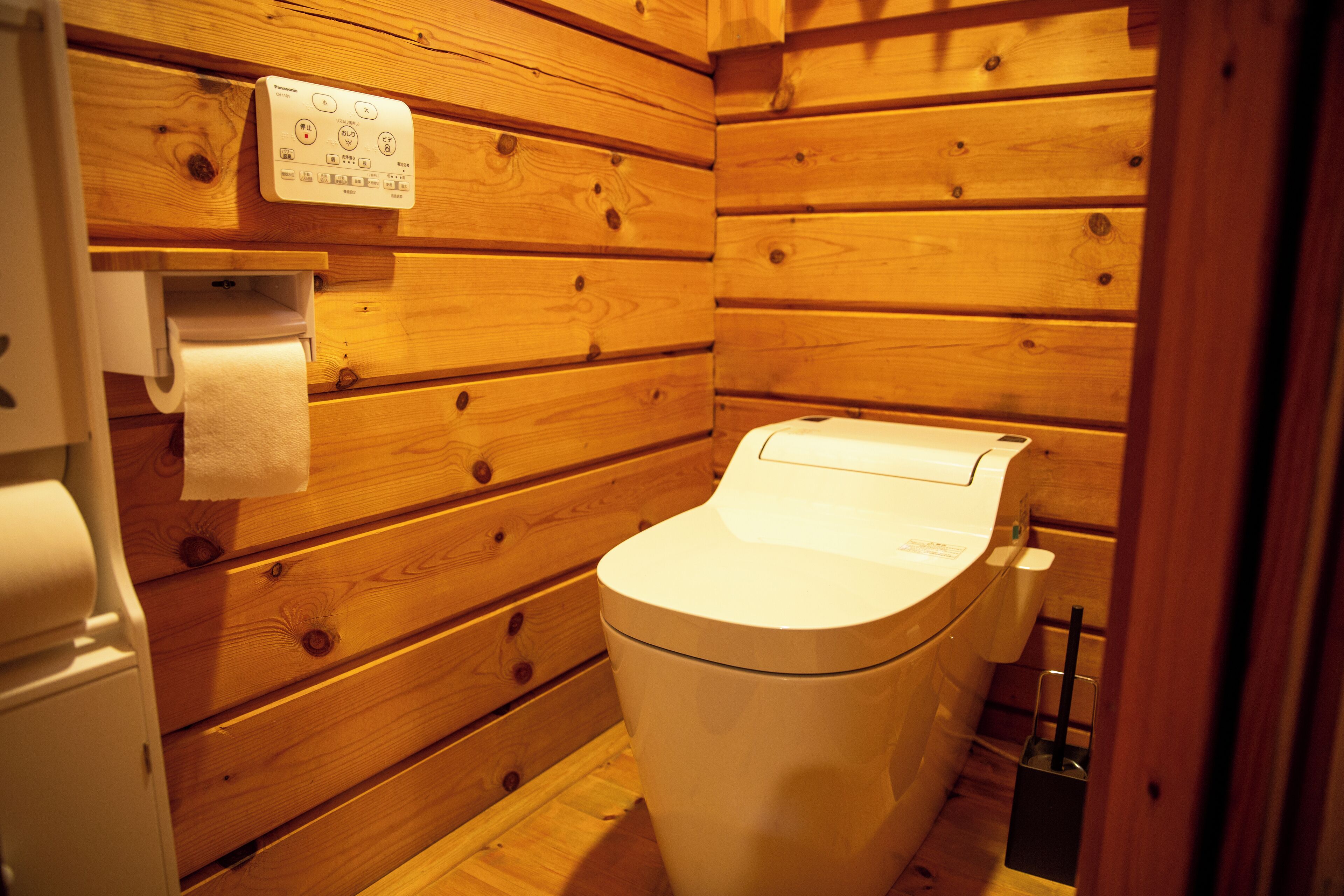 A wooden cabin-style bathroom with a toilet, a paper towel dispenser, and a small trash bin.