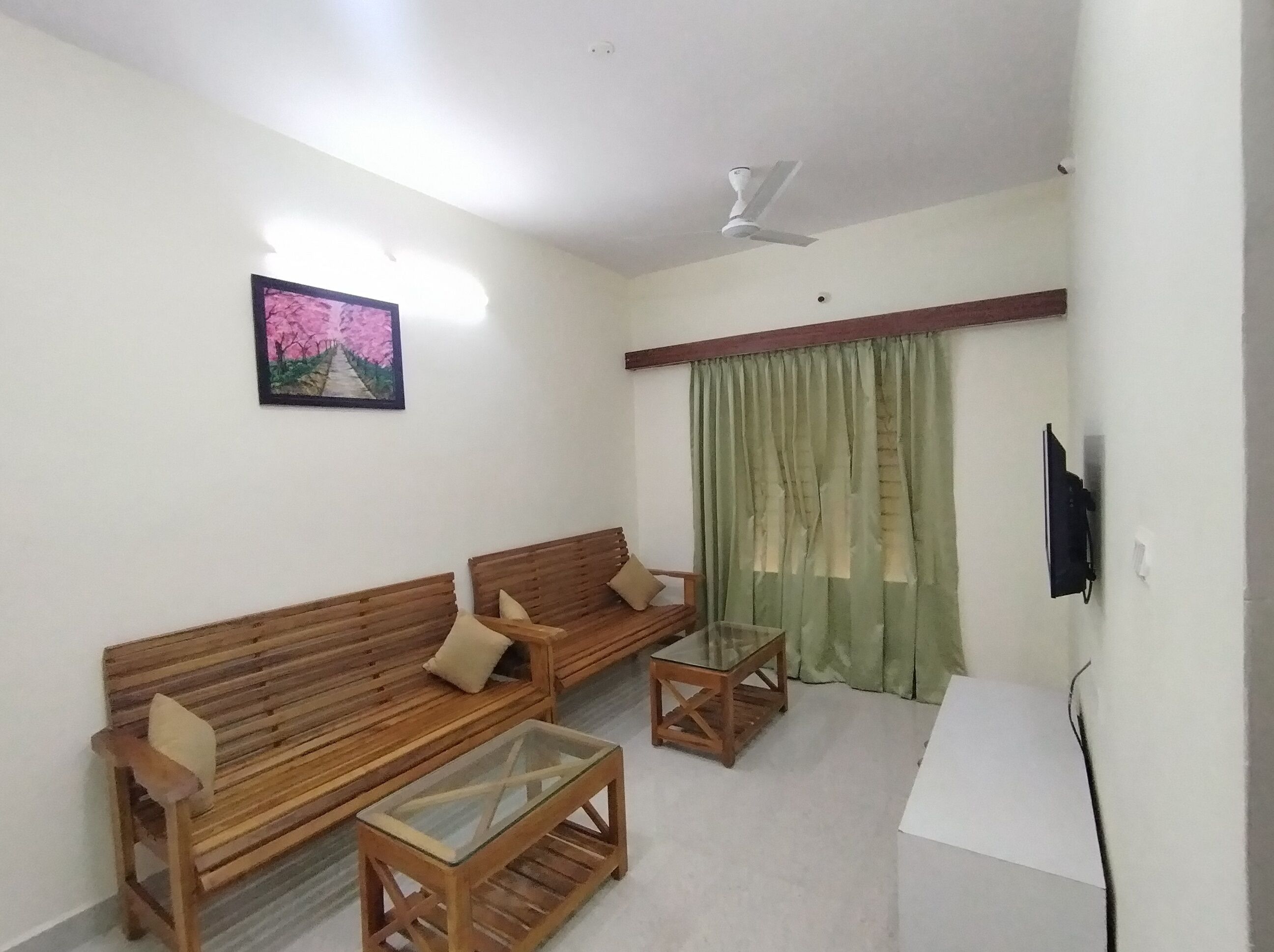 Uma sala de estar com dois sofás de madeira, uma mesa de centro de vidro, um ventilador de teto, um quadro na parede e uma cortina verde.