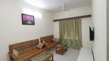 Uma sala de estar com dois sofás de madeira, uma mesa de centro de vidro, um ventilador de teto, um quadro na parede e uma cortina verde.