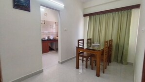 A dining area with a glass table and wooden chairs, a kitchen in the background, and a green curtain over a window. - CTS Residency (Tiruchirappalli)